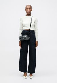 Weißer gestreifter Strick-Crop-Pullover, dunkle weiter geschnittene Jeans und eine schwarze verzierten Handtasche mit Schultergurt; weiße spitze Absatzschuhe.
