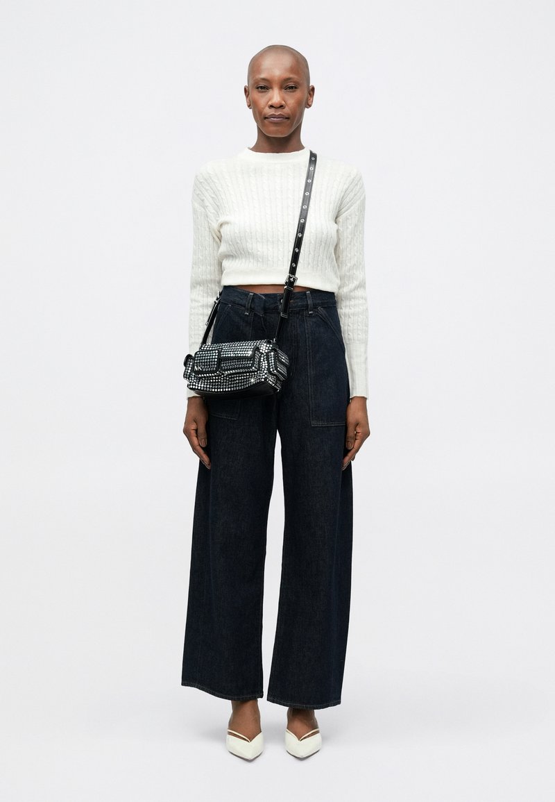 Weißer gestreifter Strick-Crop-Pullover, dunkle weiter geschnittene Jeans und eine schwarze verzierten Handtasche mit Schultergurt; weiße spitze Absatzschuhe.