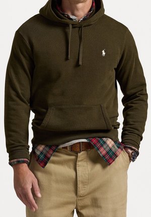 Homme portant un sweat à capuche vert olive par-dessus une chemise à carreaux rouges et verts, pantalon beige, ceinture en cuir marron et montre-bracelet au cadran sombre.