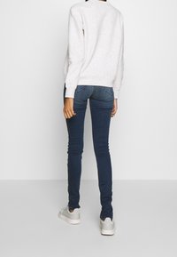 Sudadera de color gris claro con manchas, jeans ajustados azules y zapatillas grises. La sudadera tiene un cuello redondo y puños acanalados, los jeans tienen bolsillos traseros.