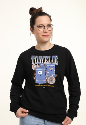 Kobieta w okularach i z włosami upiętymi w kok nosi czarną bluzę z animowanymi ręcznikami z twarzami i napisem „Towelie: Nie mam pojęcia, co się dzieje.”