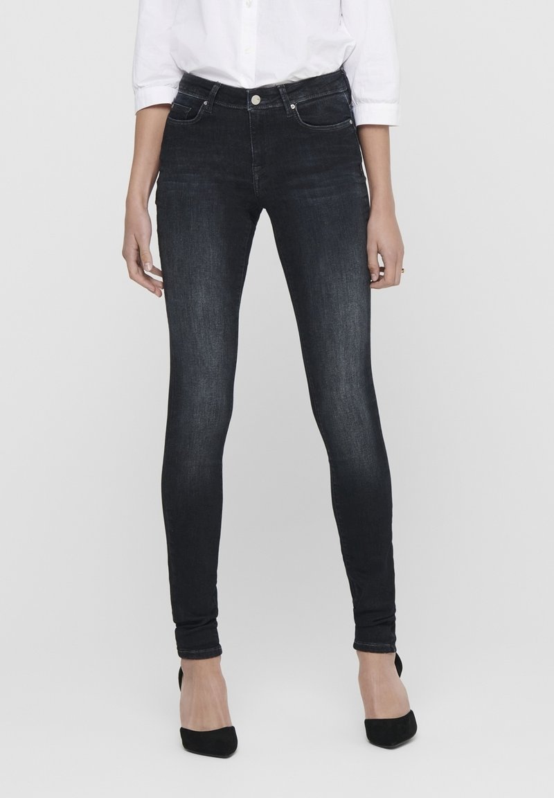 Femme portant un jean noir ajusté, une chemise blanche boutonnée et des chaussures à talons noires, se tenant devant un fond gris uni.