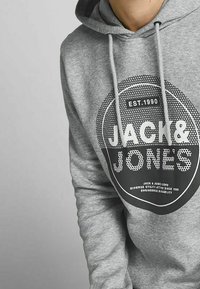 Felpa grigia con cappuccio caratterizzata da un logo circolare con il testo "JACK & JONES" e "EST. 1990" in nero a contrasto, progettata con coulisse e una finitura testurizzata.