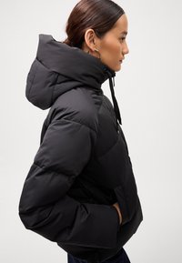 Black puffer jacket with a fitted hood, textured quilted pattern, front zipper, and drawstring closure. Features large side pockets.