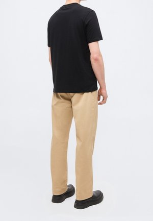Personne portant un t-shirt noir uni à manches courtes, un pantalon beige et des chaussures noires, debout de dos sur un fond blanc.