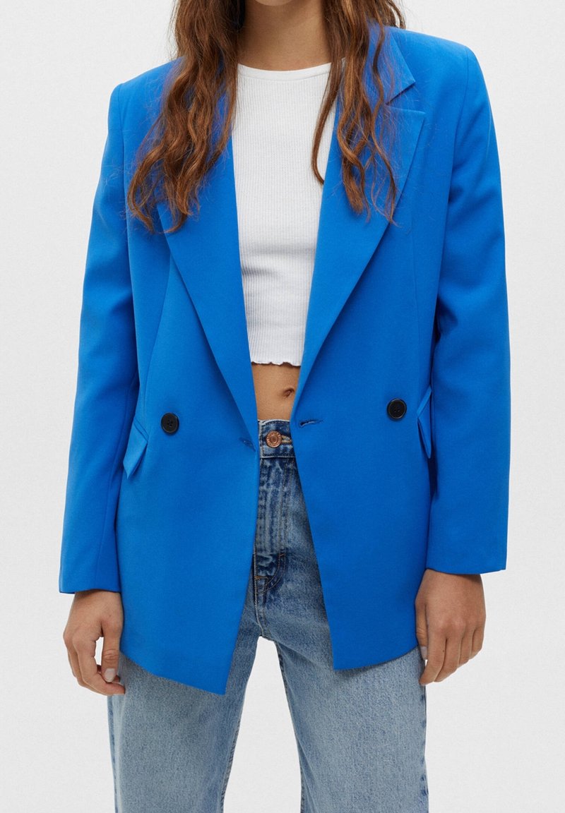 Femme portant un blazer bleu vif à double boutonnage sur un crop top blanc et un jean bleu clair, debout devant un fond uni.