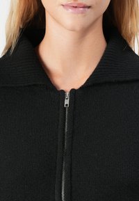 Nærbilde av en persons hals og nedre ansikt som har på seg en svart teksturert zip-up-plagg med brettet krage.