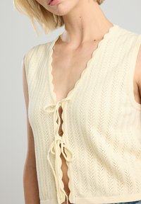 Knit sleeveless top in cream, featuring a scalloped neckline, front tie closure, and textured vertical patterns. Soft, lightweight material.