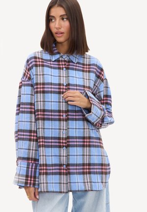 Jeune femme aux cheveux bruns raides portant une chemise ample à carreaux bleu, rose et noir et un jean bleu clair, regardant sur le côté.