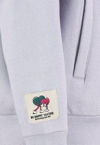 Lichtpaarse sweatshirt met een stoffen patch waarop cartoon harten elkaar omarmen, met de tekst "WEES BLIJ SAMEN MET POSITIEVE VIBES."