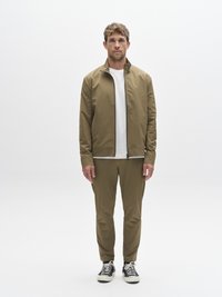 Olive rohelise värviga jakk kõrge kraega valge t-särgi peal, sobitatud kitsenevate pükstega ja mustade tossudega, millel on valged tallad.