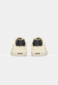 Baskets en cuir blanc avec des accents noirs au talon, surface texturée, profil bas, et un logo "BOSS" visible sur le talon.
