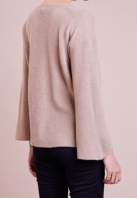 Pull en beige à côtes avec des manches longues, un col rond et une coupe décontractée. Le matériau semble doux et texturé.