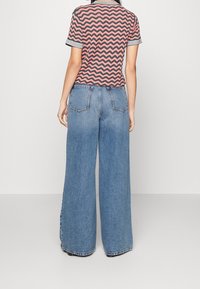 Donna che indossa jeans blu a vita alta e gamba larga, insieme a una maglietta a maniche corte con righe ondulate rosa e nere, in piedi davanti a uno sfondo bianco uniforme.