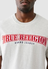 Šviesiai pilkos spalvos trumpomis rankovėmis marškinėliai su raudonai ir tamsiai mėlyna siuvinėta "TRUE RELIGION BRAND JEANS" logotipo priekyje, apvaliu iškirpimu.