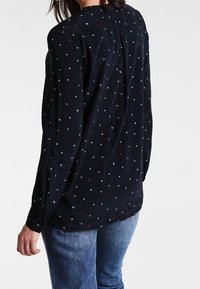 Long-sleeved black blouse featuring a scattered pattern of small white and red dots. Soft fabric, relaxed fit, and rounded hemline.