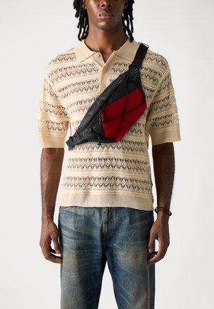 Black and red sling bag with a mesh panel, zip closure, and adjustable strap, worn crossbody over a beige patterned knit polo and blue jeans.
