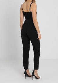 Mono negro con un cuerpo texturizado y pantalones ajustados, con tirantes finos y cierre de cremallera en la espalda; usado con tacones altos.