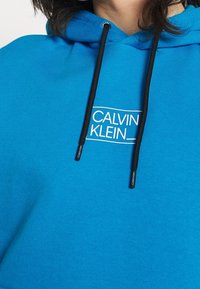 Sudadera azul hecha de una mezcla de algodón, con cordones en negro y un logo blanco de "CALVIN KLEIN" impreso en el frente.