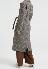 Lång, rutigt trenchcoat i svart och vitt med bälte i midjan, ventil bak och urtagna krage, kombinerad med bruna vida byxor.