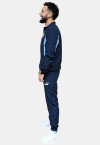 Chándal navy con detalles en azul claro, chaqueta con cremallera y puños elásticos. Combinado con zapatillas blancas y presenta el logo de Puma en los pantalones.