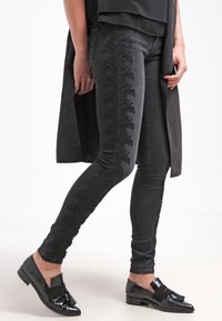 Šedé džíny skinny z denimu s propracovanou černou květinovou výšivkou po vnější straně. V kombinaci s černými lesklými mokasíny a vrstveným černým topem.