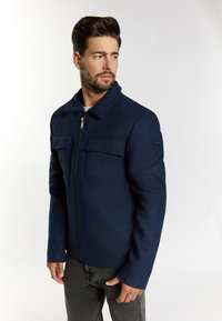 Marineblaue Wolljacke mit Reißverschluss vorne, zwei Brusttaschen und einem Kragen. Der Stoff wirkt glatt, hat einen strukturierten Schnitt und lange Ärmel.