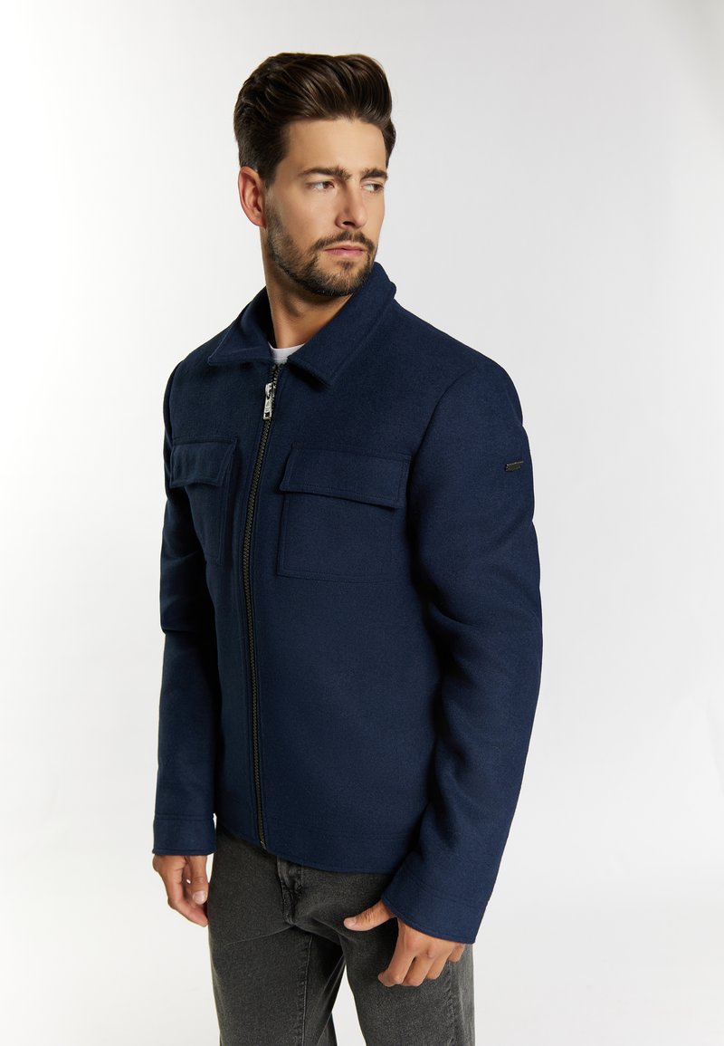 Marineblaue Wolljacke mit Reißverschluss vorne, zwei Brusttaschen und einem Kragen. Der Stoff wirkt glatt, hat einen strukturierten Schnitt und lange Ärmel.