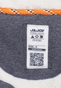 Textielflabel op een grijze stof met oranje ingeweven rand, met merknaam, verzorgingssymbolen en een QR-code tegen een witte textuur achtergrond.