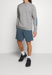 Sweat-shirt gris clair avec logo bleu, associé à un short turquoise, des baskets noires et des chaussettes blanches. Tenue de sport texturée et décontractée.