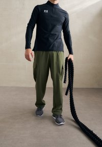 Svart långärmad sporttröja med dragkedja, olivgröna cargobyxor och grå sportskor. Håller i ett tjockt svart rep.