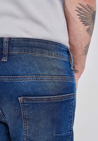 Modre denim hlače z zbledelim videzom. Značilnosti vključujejo zadnje žepke, oranžno šivanje in gladko, teksturirano blago.