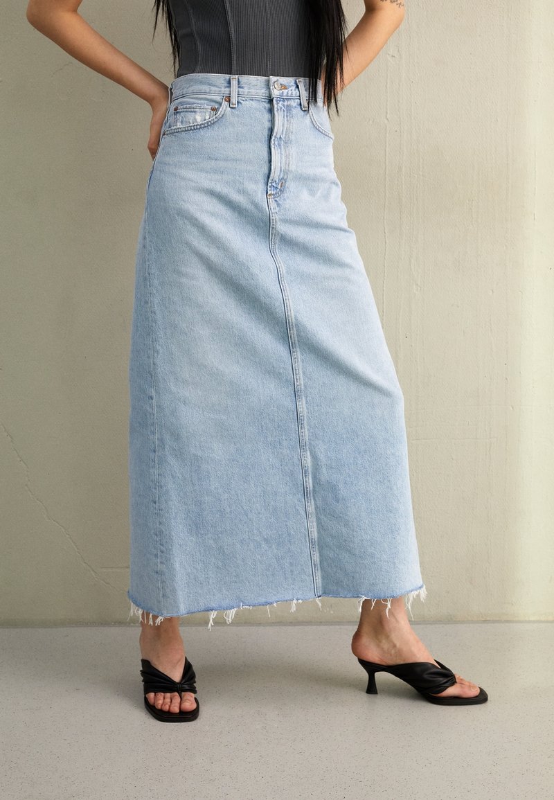 Personne portant une jupe longue en denim bleu clair à taille haute avec un ourlet effiloché et des sandales noires à talons, debout contre un mur beige.