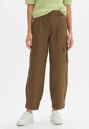 Persona con pantalones cargo marrones de talle alto con bolsillo lateral, suéter de punto verde y zapatillas blancas sobre un fondo blanco.