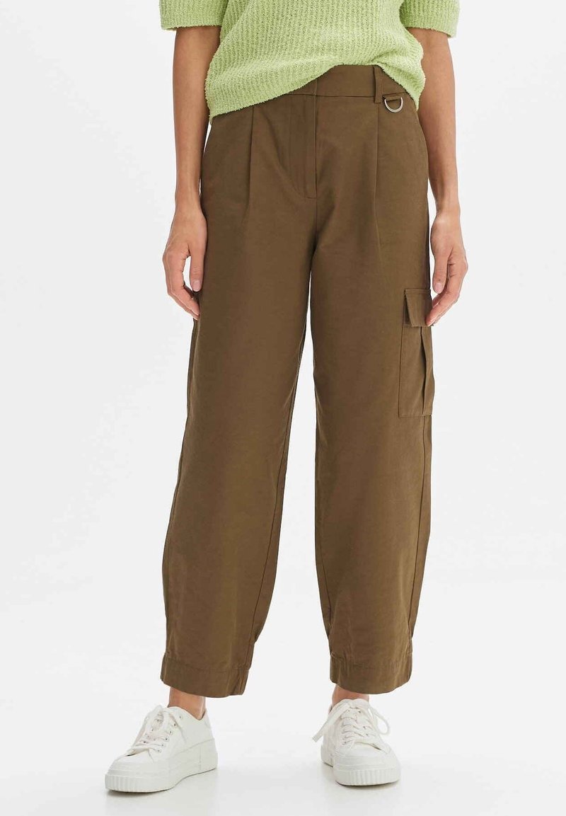 Personne portant un pantalon cargo marron taille haute avec poche latérale, un pull en maille vert et des baskets blanches sur fond blanc.