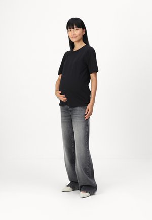 Schwarzes kurzärmeliges Umstandshirt, kombiniert mit weit geschnittenen grauen Jeans und spitzen weißen Schuhen, getragen von einer Person, die steht und ihren Bauch hält.