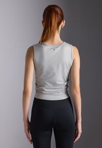 A woman with long red hair faces away, wearing a fitted gray tank top and black leggings, against a muted gray background.