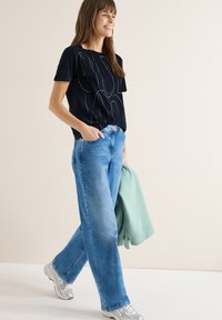 Femme marchant, portant un t-shirt graphique bleu marine, un jean bleu clair, des baskets argentées, tenant un sweat-shirt vert menthe plié dans une main.