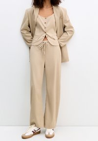 Femme portant un pantalon en maille côtelée beige, un gilet boutonné assorti, un blazer surdimensionné et des baskets blanches avec des accents marron, se tenant devant un mur blanc.