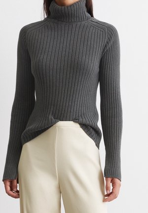 Frau trägt einen eng anliegenden dunkelgrauen, gerippten Rollkragenpullover, der in eine hoch taillierte cremefarbene Hose gesteckt ist, vor einem schlichten Hintergrund.