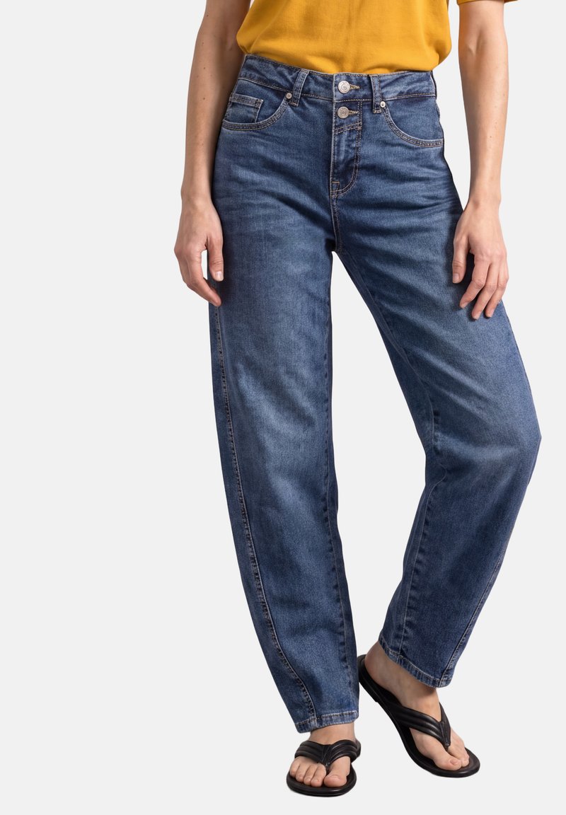 Personne portant un jean bleu taille haute avec une fermeture à double bouton, associé à des sandales noires et un haut jaune moutarde, posant avec les mains détendues.