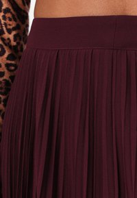 Gonna plissettata bordeaux con una cintura liscia. Realizzata in un tessuto leggero, presenta pieghe verticali che creano una texture fluida.