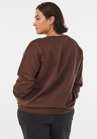 Ruda laisvo kirpimo džemperis su ilgomis rankovėmis, apvalia iškirpte ir ribbiniu apačios apvadu. Lygi tekstūra su minimaliais siūlais ir be matomų raštų.