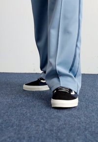 Sapatilhas baixas em preto e branco com superfície texturizada e detalhes em tecido trançado, emparelhadas com calças largas de tom azul claro sobre um carpete azul.