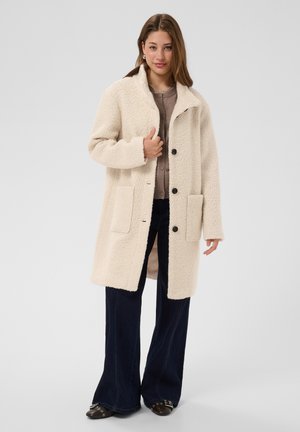 Femme portant un manteau texturé de couleur crème par-dessus une chemise marron boutonnée, un pantalon évasé foncé et des chaussures noires avec des boucles en métal.