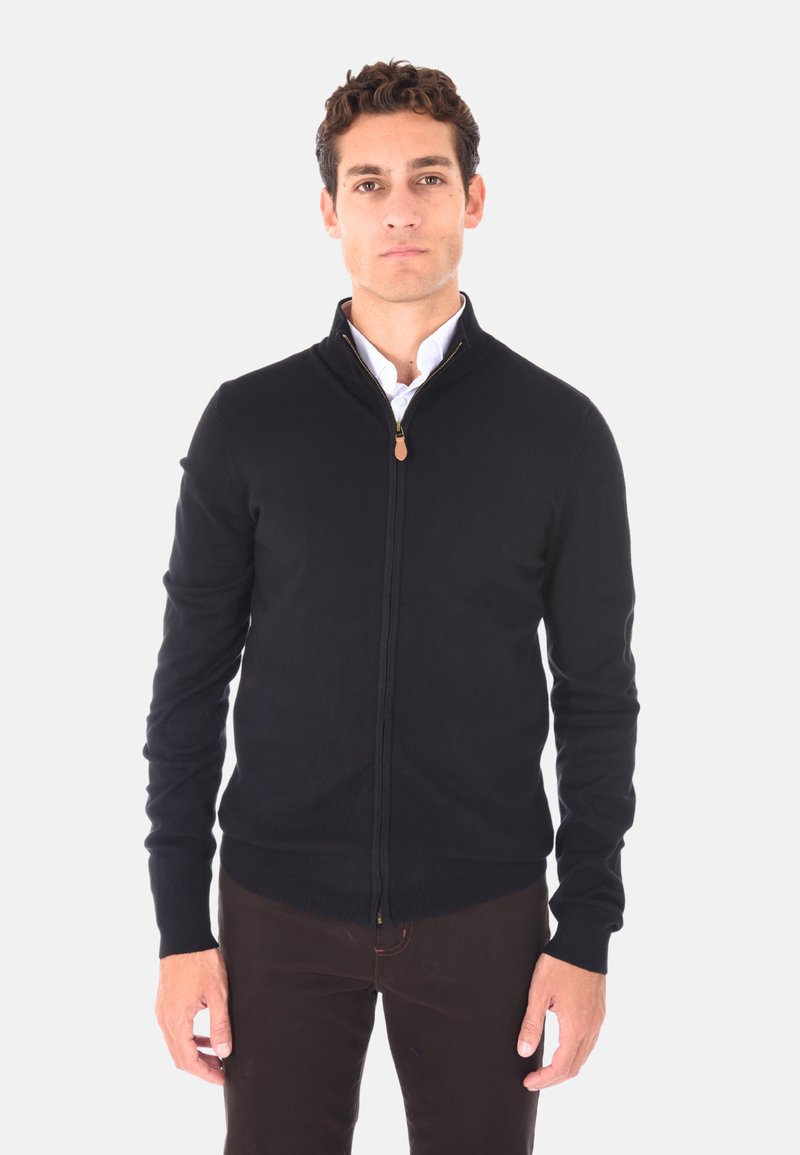 Cardigan zip-up nero in tessuto a maglia, con colletto alto, polsini a coste e una zip di colore beige. Indossato sopra una camicia bianca.