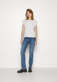 Camiseta de algodón blanca de manga corta con un pequeño logo; combinada con jeans azules y zapatos negros, de pie sobre un fondo neutro.