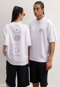 White cotton t-shirts feature a sun graphic and text on the back. The loose fit is paired with black shorts, creating a casual look.