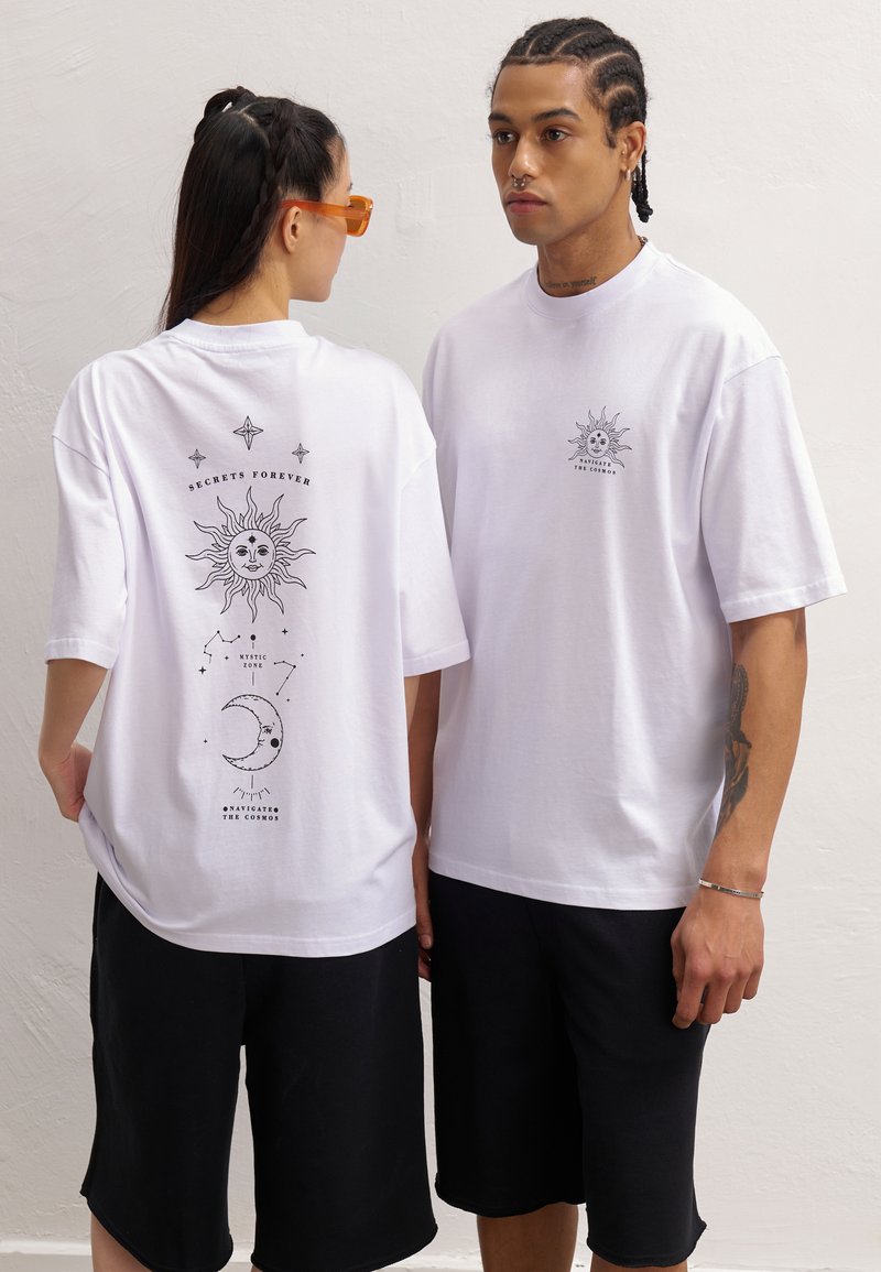 White cotton t-shirts feature a sun graphic and text on the back. The loose fit is paired with black shorts, creating a casual look.
