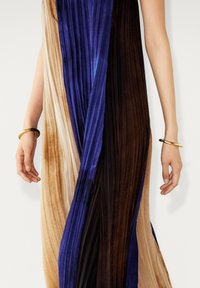 Frau trägt ein plissiertes Kleid mit vertikalen beigen, blauen und braunen Paneelen sowie goldenen Armbändern an beiden Handgelenken, vor einem schlichten weißen Hintergrund.